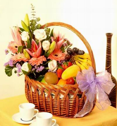 Fruit and Flower basket of 3 Lilies 6 light pink Roses, 4 pink Gerbera Daisies, purple Stock and Greenery with 6 Strawberries, 5 Bananas, 1 Mango, 1 Orange, 1 Kiwi, 1 green Apple, and purple Grapes.