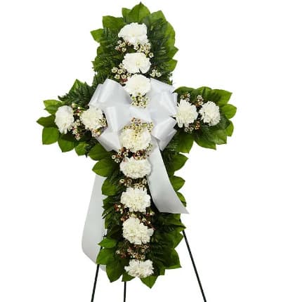 This white flower standing cross is a thoughtful tribute of sympathy. Created by a florist with white carnations, white waxflower and seasonal greens, in a shape of a cross. Delivered on an easel with a white decorative bow. Measures 18